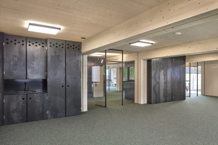 Gruppenraum mit Blick in den Korridor, mit Glas abgetrennt. Sichtbare Holzbalken und Deckenplatten. Grüner Teppich.