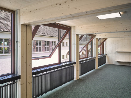 Sicht von einem Schulzimmer Richtung Flügelfenster, die halb geöffnet sind. Holzbalken und Rückwand aus Holz, Teppich