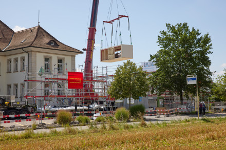 Sicht auf die Baustelle: Ein Holzbaumodul hängt am Kran.