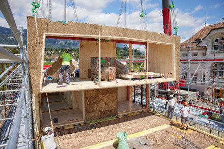 Sicht auf die Baustelle: Ein Holzbaumodul hängt am Kran und schwebt ca. 3 Meter über dem Bestimmungsort.