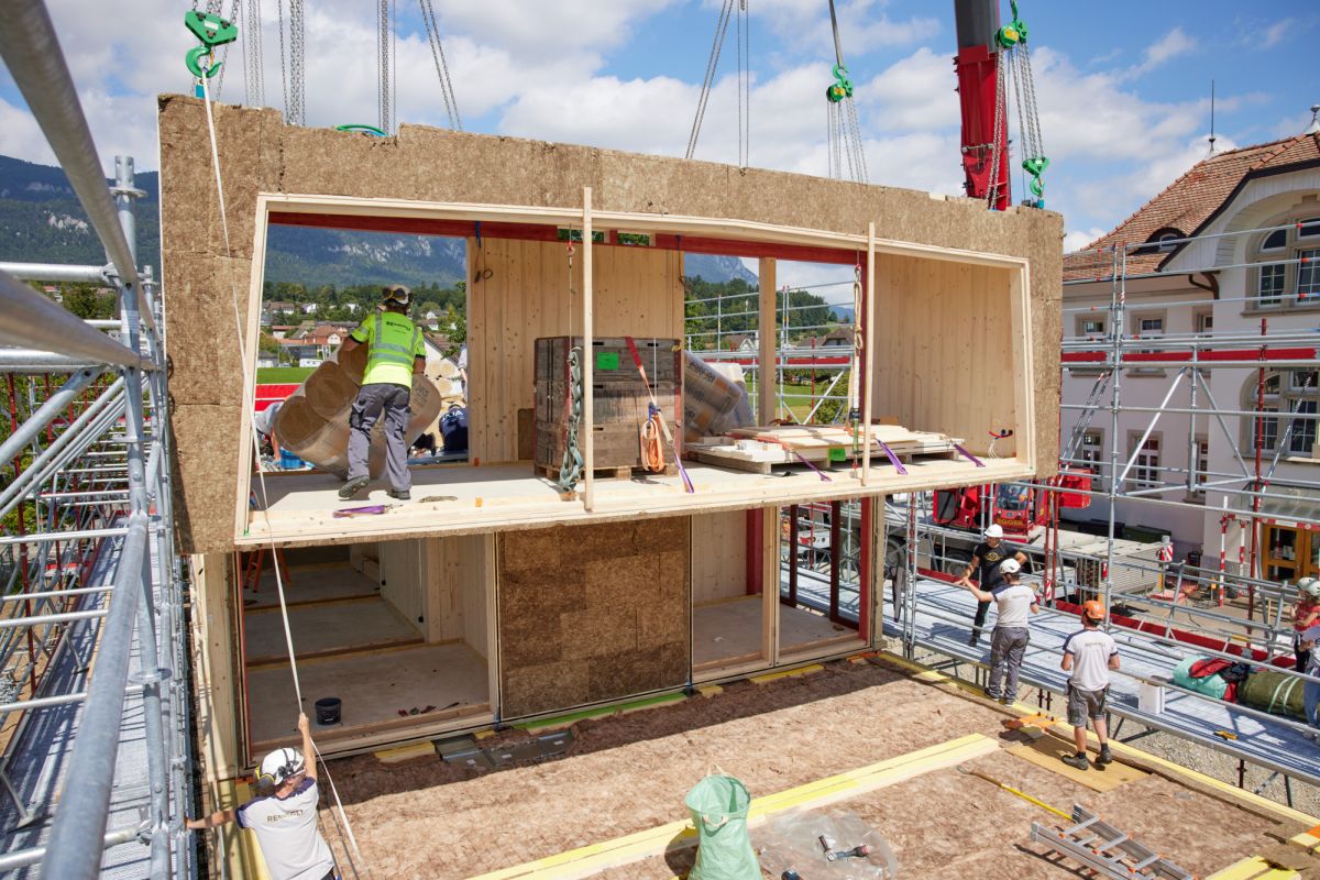 Sicht auf die Baustelle: Ein Holzbaumodul hängt am Kran und schwebt ca. 3 Meter über dem Bestimmungsort.