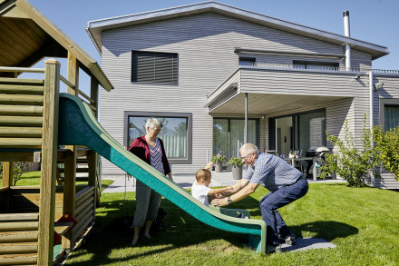 Bauherrschaft mit Enkel auf dem Spielplatz mit gedecktem Balkon und Renggli-Haus im Hintergrund