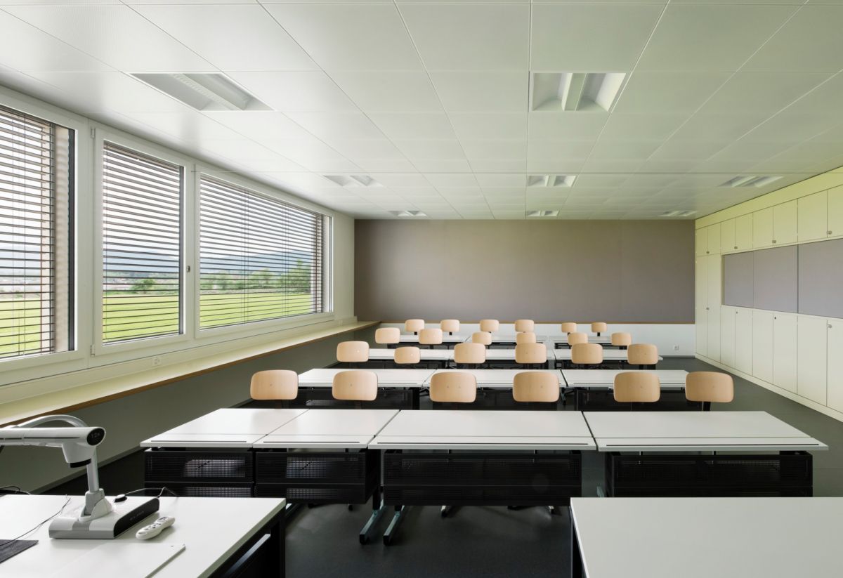 Schulzimmer mit grossen Fenstern und heruntergelassenen, quer gestellten Lamellenstoren zur Beschattung und damit indirektem Tageslicht sowie weiterhin der Aussicht.
