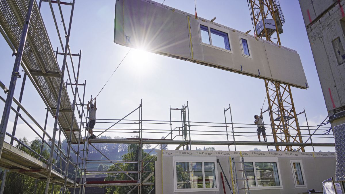 Auf einer Baustelle hängt ein Wandelement am Kran und wird von Zimmerleuten versetzt.