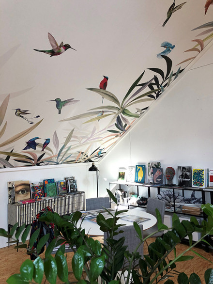 Un coin de l’atelier avec de profondes étagères, une table, des plantes et, sur la pente du toit, un papier peint orné d’élégantes plantes et d’oiseaux.