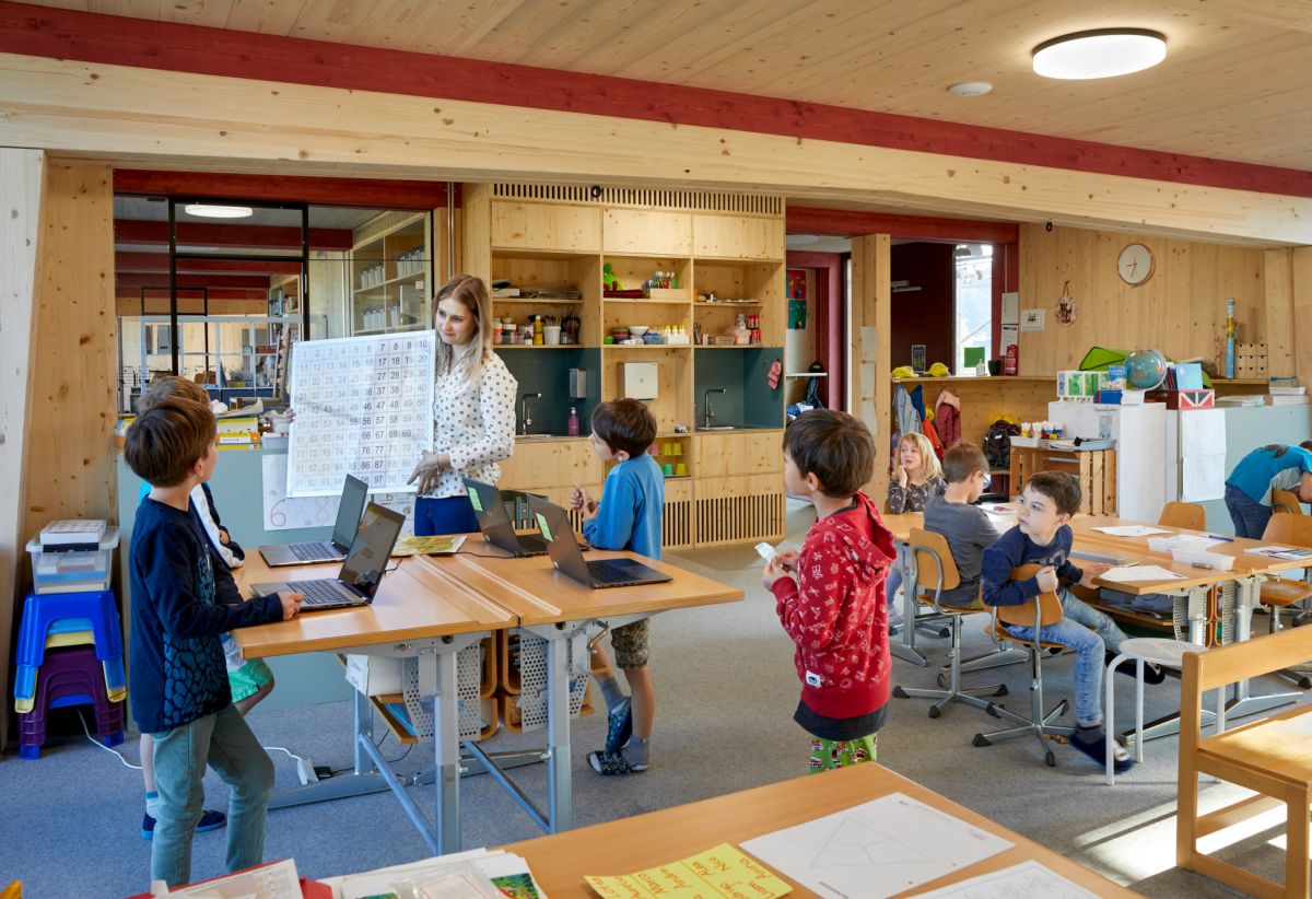 Des élèves du primaire écoutent leur enseignante dans une salle de classe avec beaucoup de bois visible.