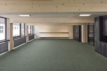 Salle de classe avec moquette, poutres et plafond en bois apparents, radiateurs et grandes fenêtres