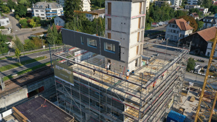 Chantier avec la cage d’escalier en acier et béton. Echafaudage pour le montage des éléments de parois et de plafonds. Une grue hisse un long élément de paroi.