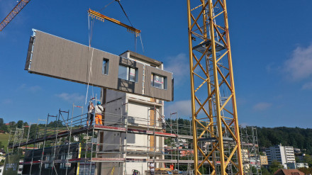 Un élément de paroi avec façade et fenêtres flotte dans l’air sur un chantier.