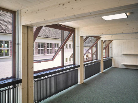 Vista da un'aula in direzione delle finestre a mezza anta aperte. Travi di legno e parete di fondo in legno, moquette
