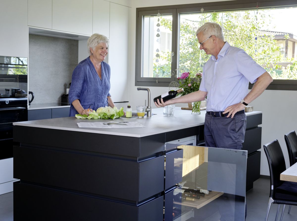 Couple debout dans une cuisine ouverte avec îlot de cuisine et fenêtre à mi-hauteur en arrière-plan, au premier plan un réfrigérateur à vin ouvert